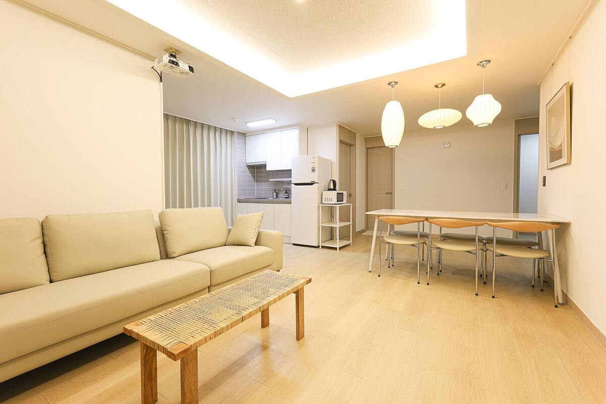 Un salon moderne comprenant un canapé, une table basse en bois, une table à manger avec des chaises, un coin cuisine et des luminaires suspendus.