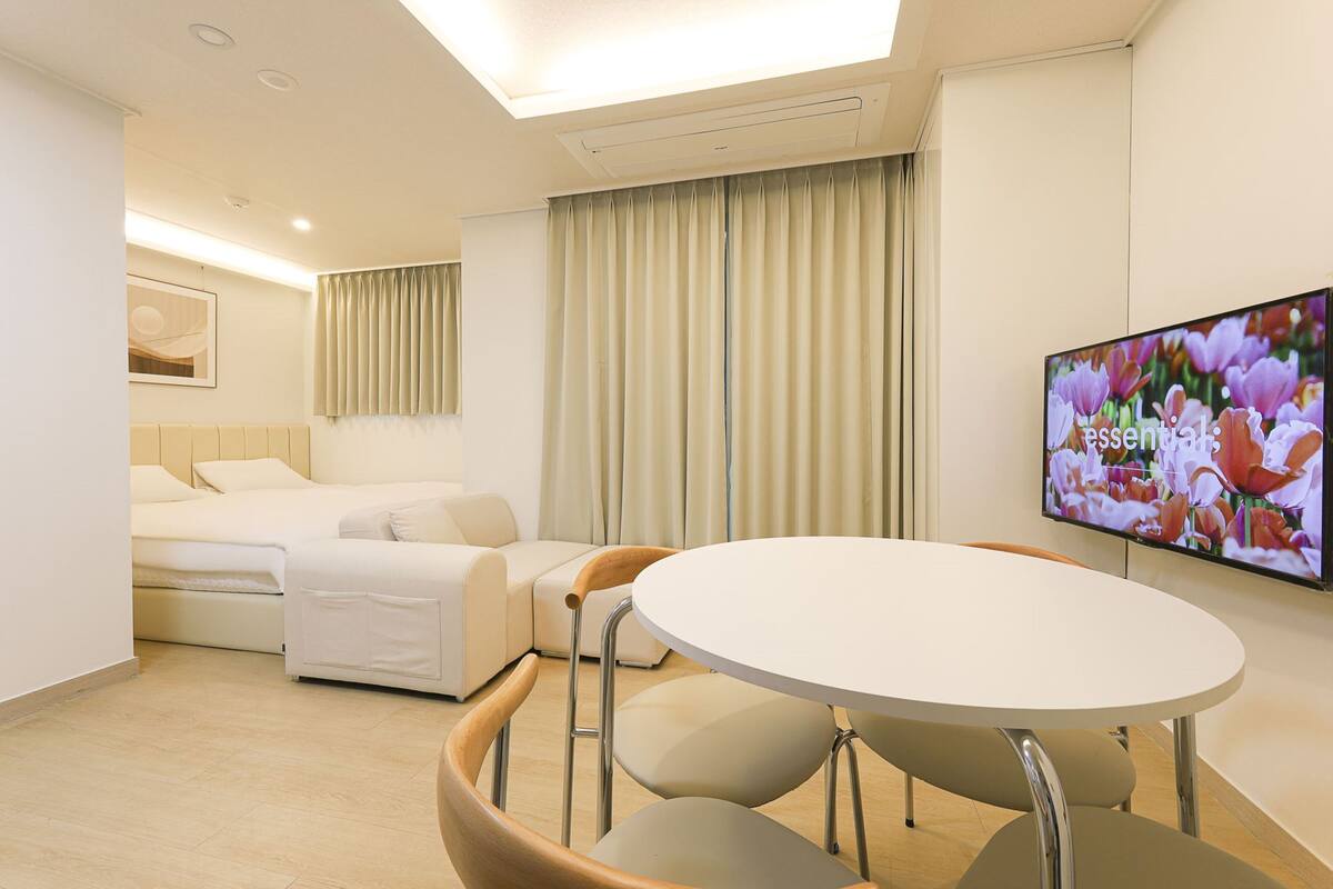 Une chambre d’hôtel moderne équipée d’un lit, d’un canapé, d’une table à manger avec des chaises, d’un téléviseur à écran plat et d’une grande fenêtre avec des rideaux.