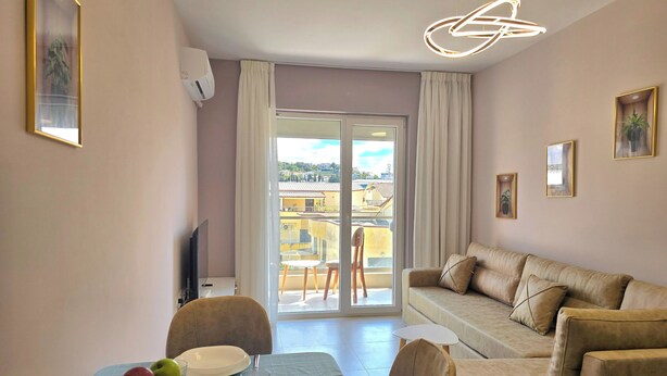 A modern living room with a sofa, dining table, and chairs. There is a balcony with a view of buildings and a cityscape. - Happy Stay Apartment By The Sea 094 (Golem)