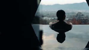 Una persona seduta su una superficie riflettente, con una città e montagne sullo sfondo.