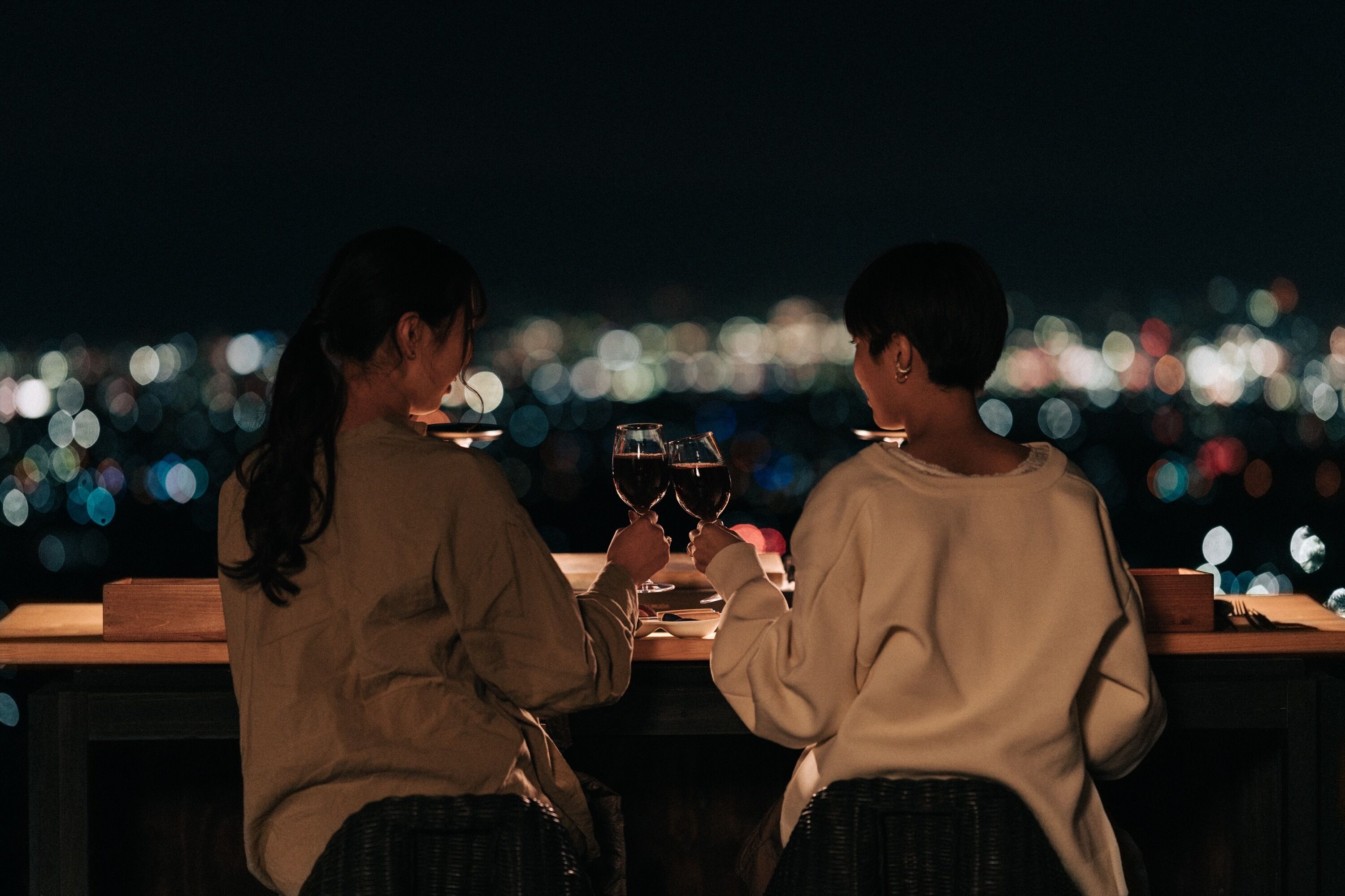 Two people sitting at a table with wine glasses, overlooking a cityscape at night.