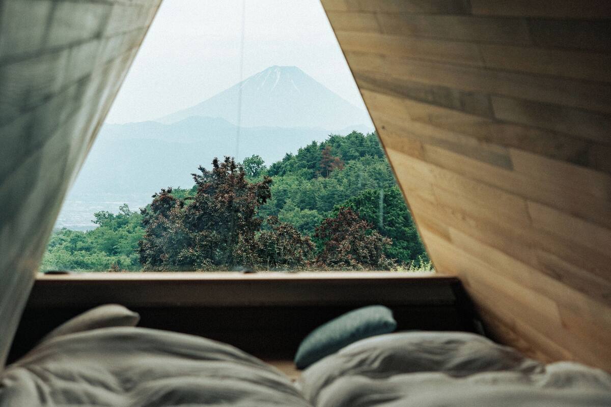 A room with a sloped ceiling and a bed with pillows. A window reveals a view of a mountain and greenery.