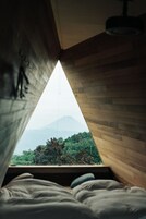 Quarto com teto inclinado e cama com almofadas. Uma janela mostra vista para montanha e vegetação.