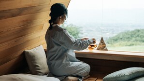 Una persona seduta vicino a una finestra con vista su un paesaggio verde, pareti di legno e un letto con cuscini.