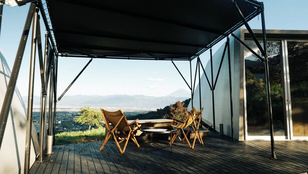 Una terrazza esterna con tavolo e sedie, vista su una città e montagne, e un edificio moderno con ampie finestre.