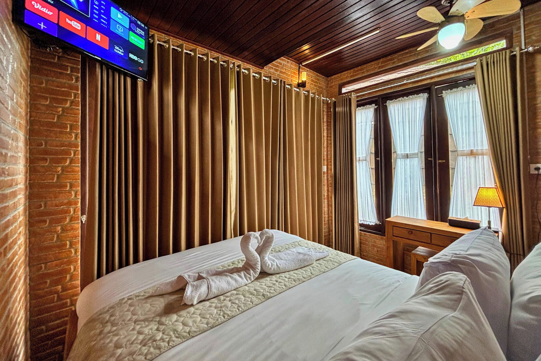 Une chambre d’hôtel avec un mur en briques, un lit agrémenté d’une serviette en forme de cygne, un plafond en bois et un téléviseur fixé au mur.