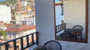 A balcony with a table and chairs, a potted plant, and a view of a cityscape with buildings and a church tower. - Ocean view apartment in downtown Vallarta a few steps from the Malecon (Puerto Vallarta)