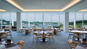 A spacious dining area with large windows offering a view of a body of water and distant hills. Tables are arranged for dining, and the floor has a patterned tile design. - Sagewood Yeosu Gyeongdo Resort (Yeosu)