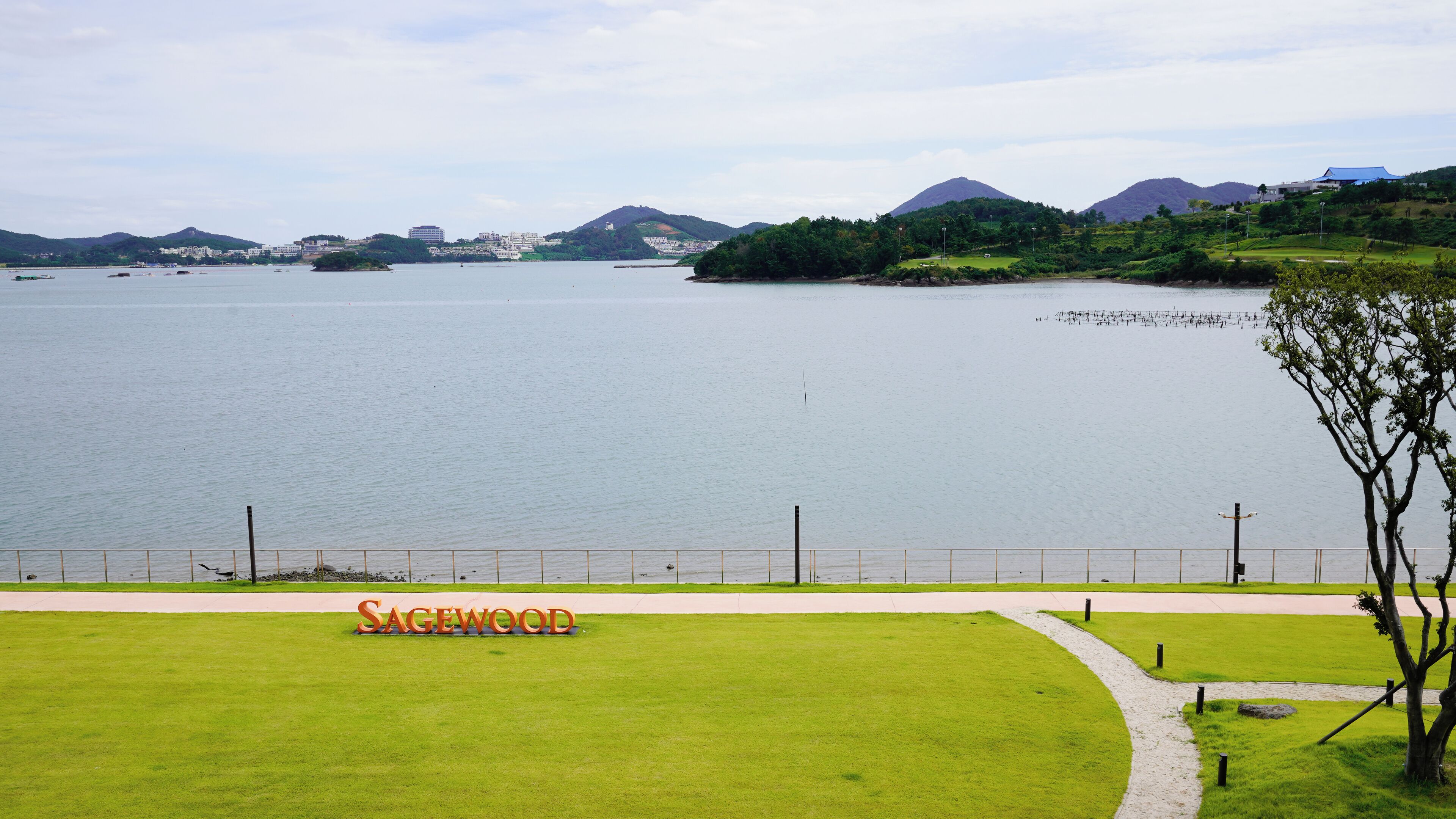 A lake with a sign reading "Sagewood" in the foreground, surrounded by green hills and buildings in the background.