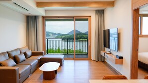 A modern hotel room with a sofa, a coffee table, a TV, and a balcony with a view of mountains and a lake. - Sagewood Yeosu Gyeongdo Resort (Yeosu)