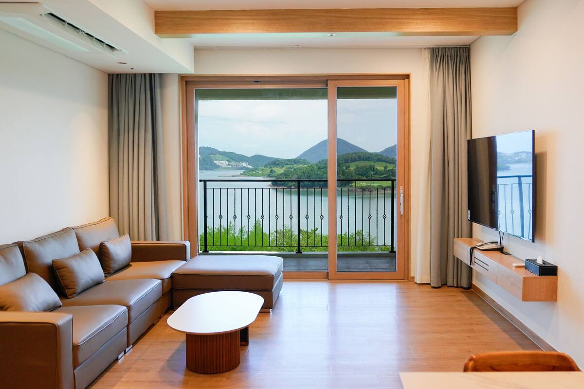 A modern hotel room with a sofa, a coffee table, a TV, and a balcony with a view of mountains and a lake.
