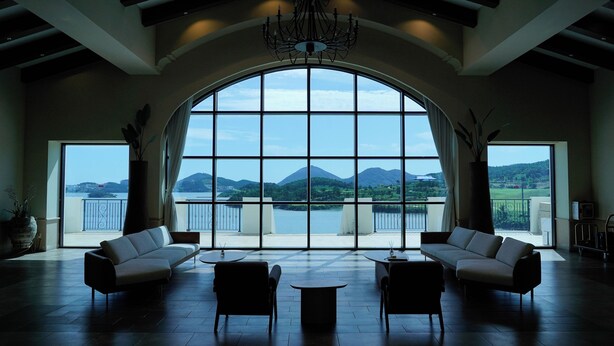 A spacious room with a large arched window offering a view of a lake and mountains. There are two sofas and a small table in the center. - Sagewood Yeosu Gyeongdo Resort (Yeosu)