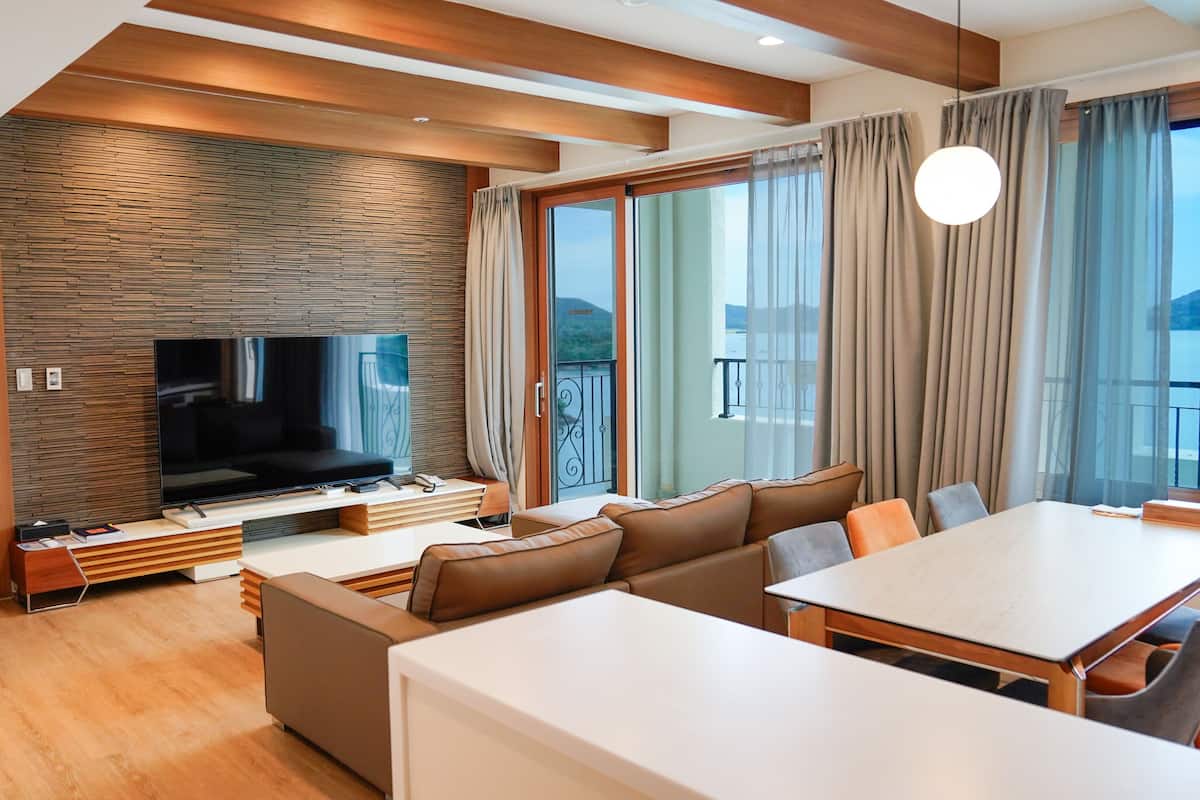 A modern hotel room with a large flat-screen TV, a sofa, a dining table, and a balcony with curtains.
