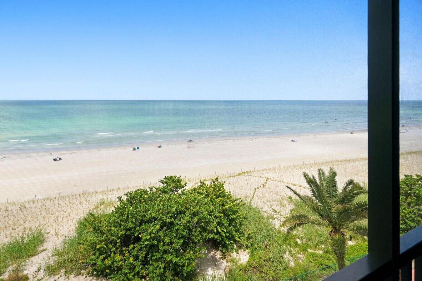 A beach view from a window overlooking a sandy beach with clear water, some vegetation in the foreground, and a clear sky.