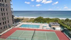 Uma quadra de tênis com piscina, vista para a praia e um edifício ao fundo.