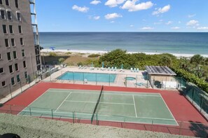 A tennis court with a pool, beach view, and a building in the background. - Beachfront Condo w/ Stunning Gulf Views | Reflections On The Gulf 504 (Indian Rocks Beach)