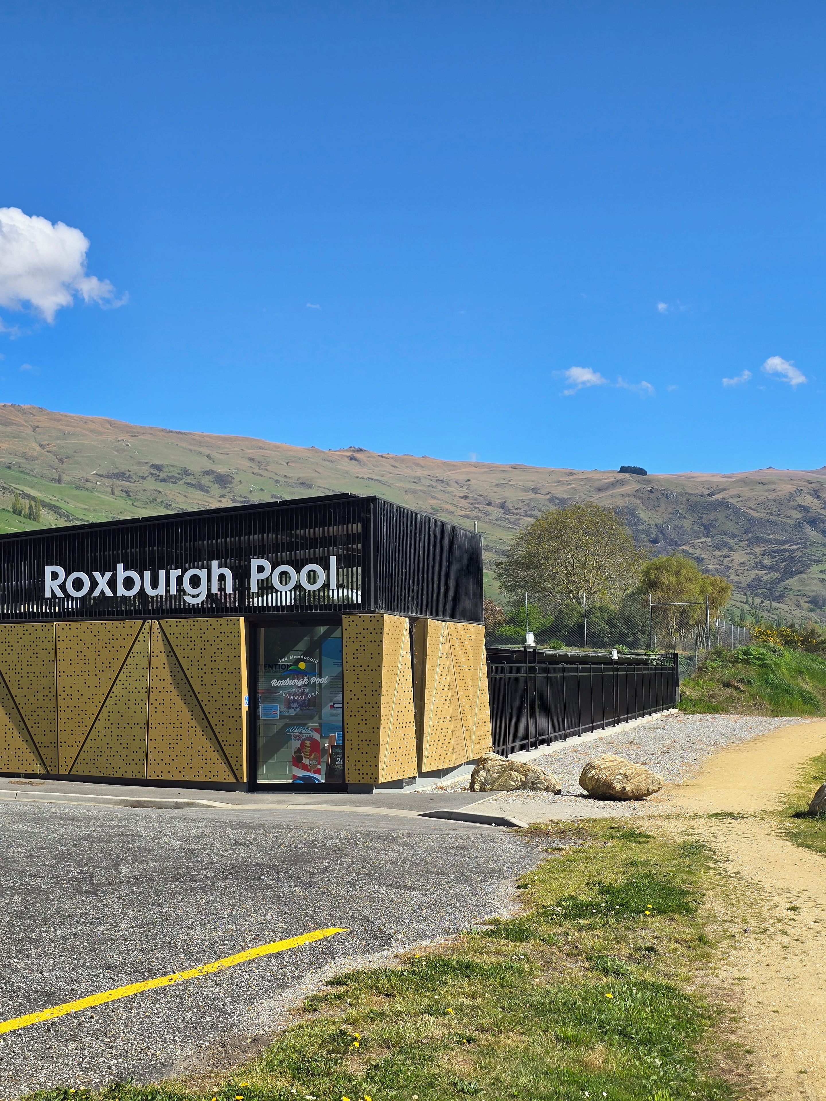 Un bâtiment portant l’inscription « Roxburgh Pool », avec un ciel dégagé et des collines en arrière-plan.