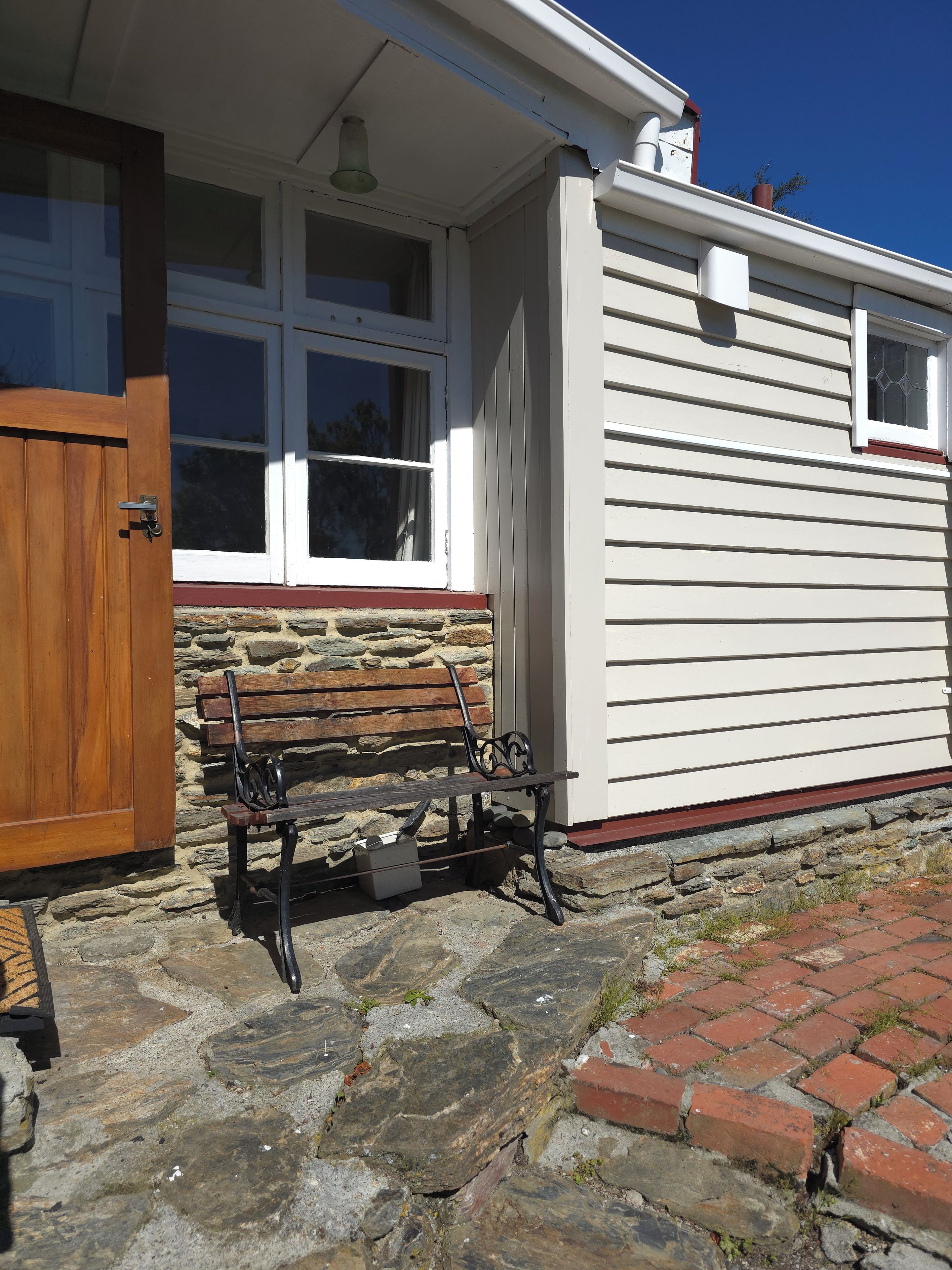 A stone-paved entrance with a wooden bench, a white door, and a window with a stone wall foundation.
