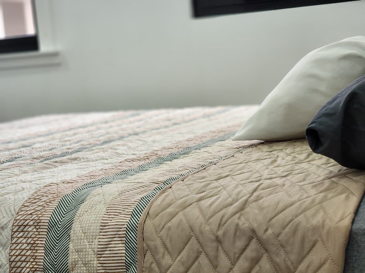 Een bed met een gewatteerde spreidek en een kussen, een raam op de achtergrond en een gedeeltelijk zicht op een muur.