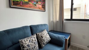 En blå sofa med mønstrede puder, et indrammet billede over og et vindue med gardiner.