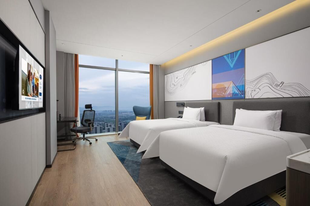 A hotel room with two beds, a desk, and a chair. There is a large window with a city view.