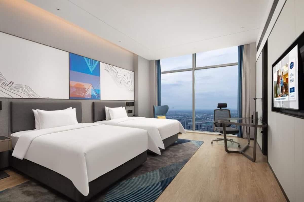 Ein modernes Hotelzimmer mit zwei Betten, einem Schreibtisch und einem großen Fenster mit Blick auf die Stadt.