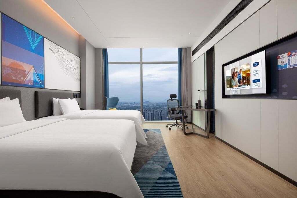 A modern hotel room with two beds, a large window with a city view, a desk, and a TV screen displaying a message.