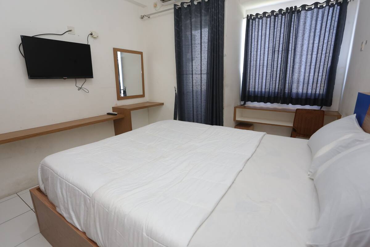 A bedroom with a bed, a mounted TV, a mirror, a window with curtains, and a bedside table.