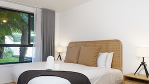 Habitación de hotel con una cama grande, una mesita de noche con lámpara y vistas a palmeras a través de la ventana.