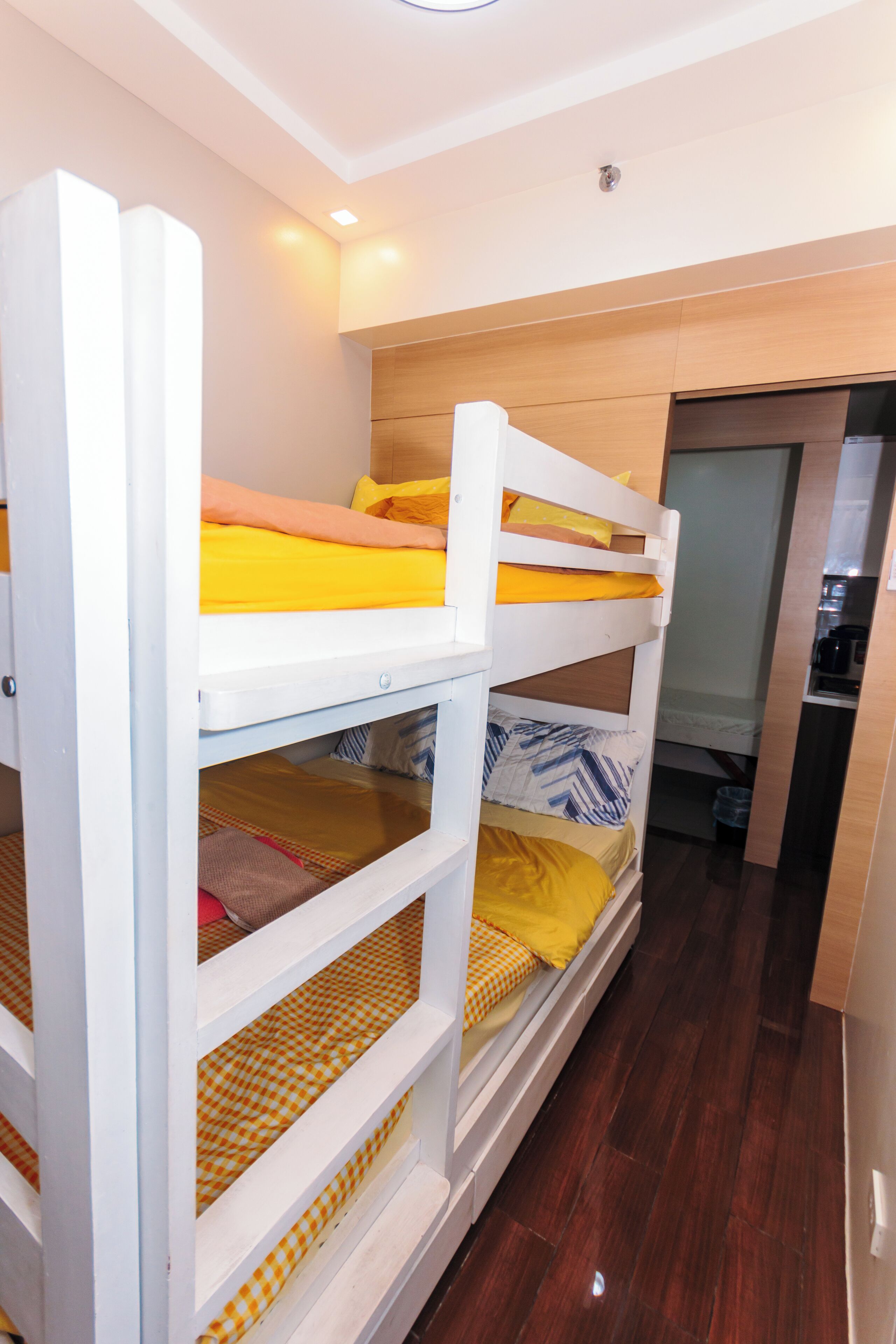 A bunk bed room with wooden flooring, a kitchen area, and a door leading to another room.