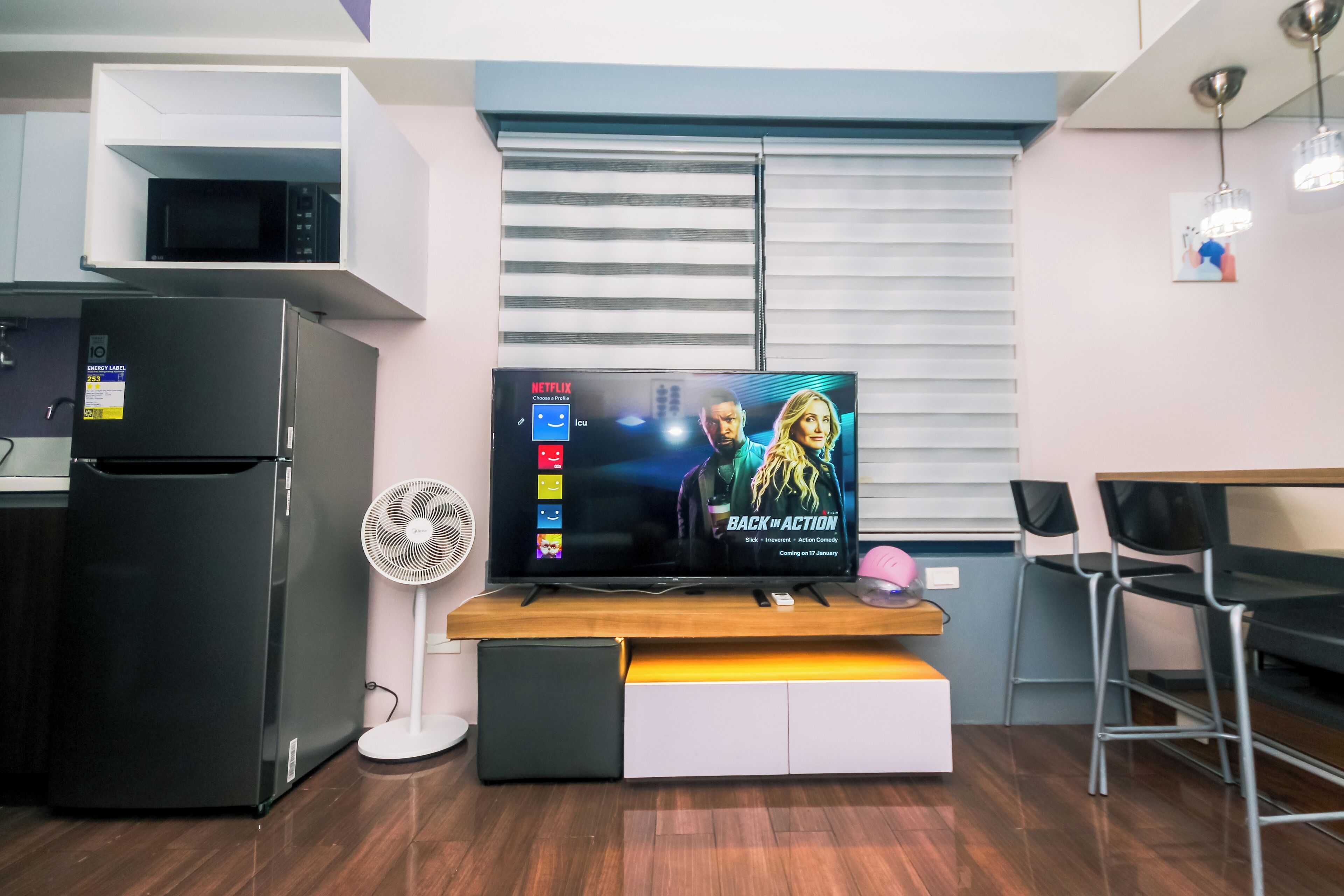 Un espacio habitable compacto con una televisión de pantalla plana sobre un soporte de madera, un refrigerador negro, un ventilador blanco y una zona de comedor con mesa y sillas.