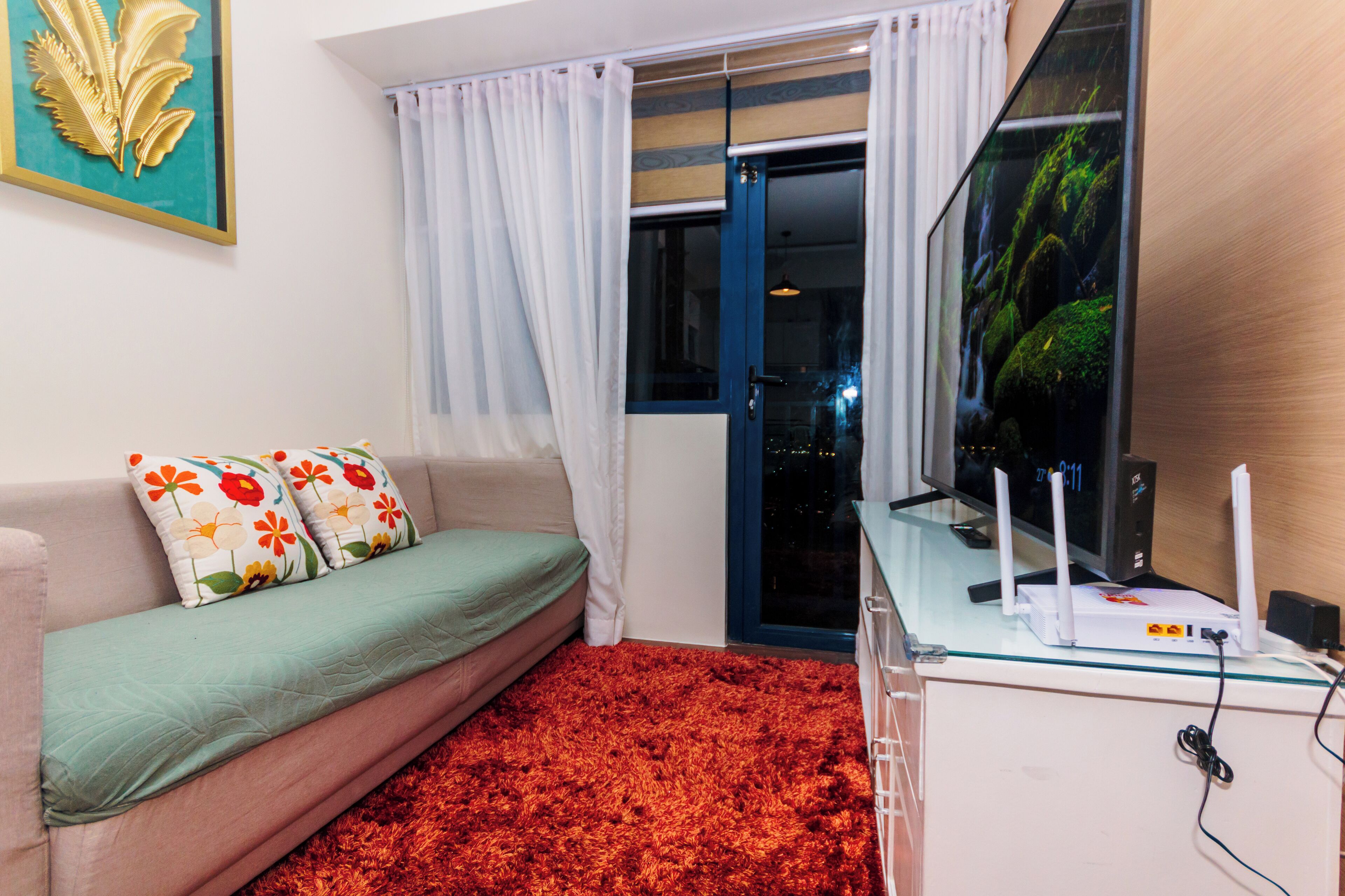 A room with a sofa, a glass table with electronic devices, a red carpet, and a view of a cityscape through a glass door.