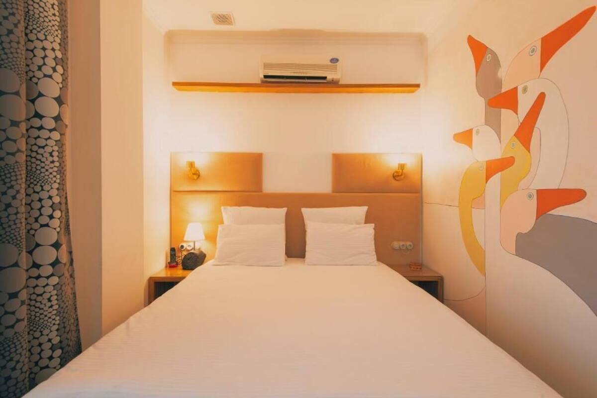 A hotel room with a bed, two bedside tables, a wall-mounted air conditioner, and a decorative wall piece.