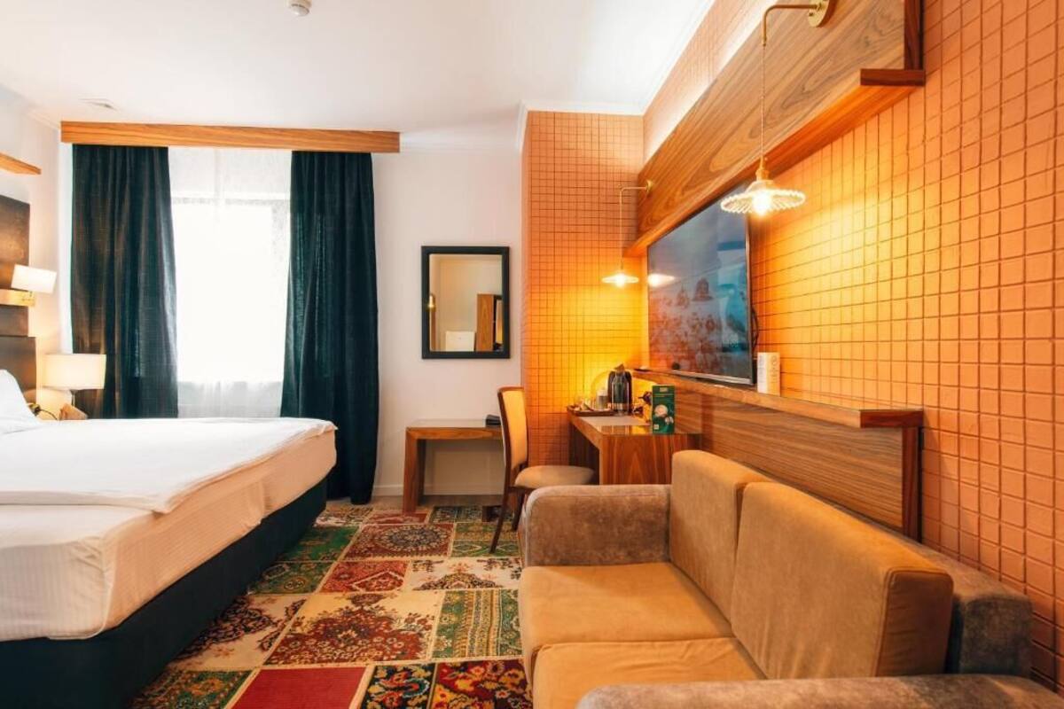 A hotel room with a bed, a sofa, a small table with a lamp, and a mirror on the wall.