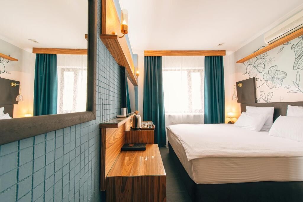 A hotel room with a bed, a wooden desk, a mirror, and a wall with a floral pattern.