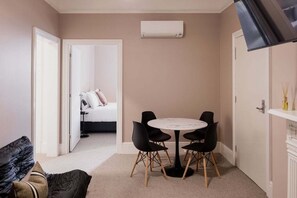 A compact room with a round table and four chairs, a bed in the background, and an air conditioning unit on the wall. - Palfreymans Arcade | Central NoHo 2 Bed Apartment - 1 (Hobart City)