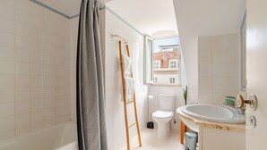 A bathroom with a bathtub, toilet, sink, and a window with a view of buildings. - Cosy Loft Heart Of The City! (Lisbon)
