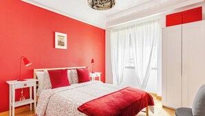 A bedroom with a red wall, a bed with floral bedding, a red throw blanket, a white wardrobe, and a desk with a lamp. - Top Location Beautiful Apartment! (Lisbon)