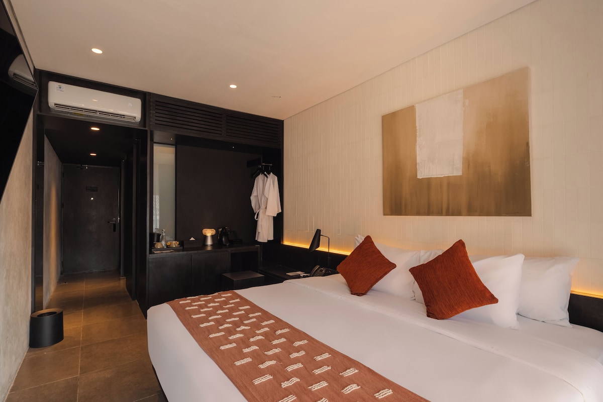 A hotel room with a bed, a desk, a mirror, and an air conditioning unit.