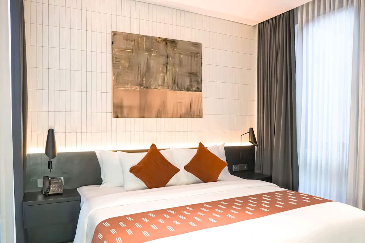 Una camera d'albergo moderna con un letto grande, un comodino, una lampada da tavolo, un quadro astratto appeso alla parete e una finestra con tende.