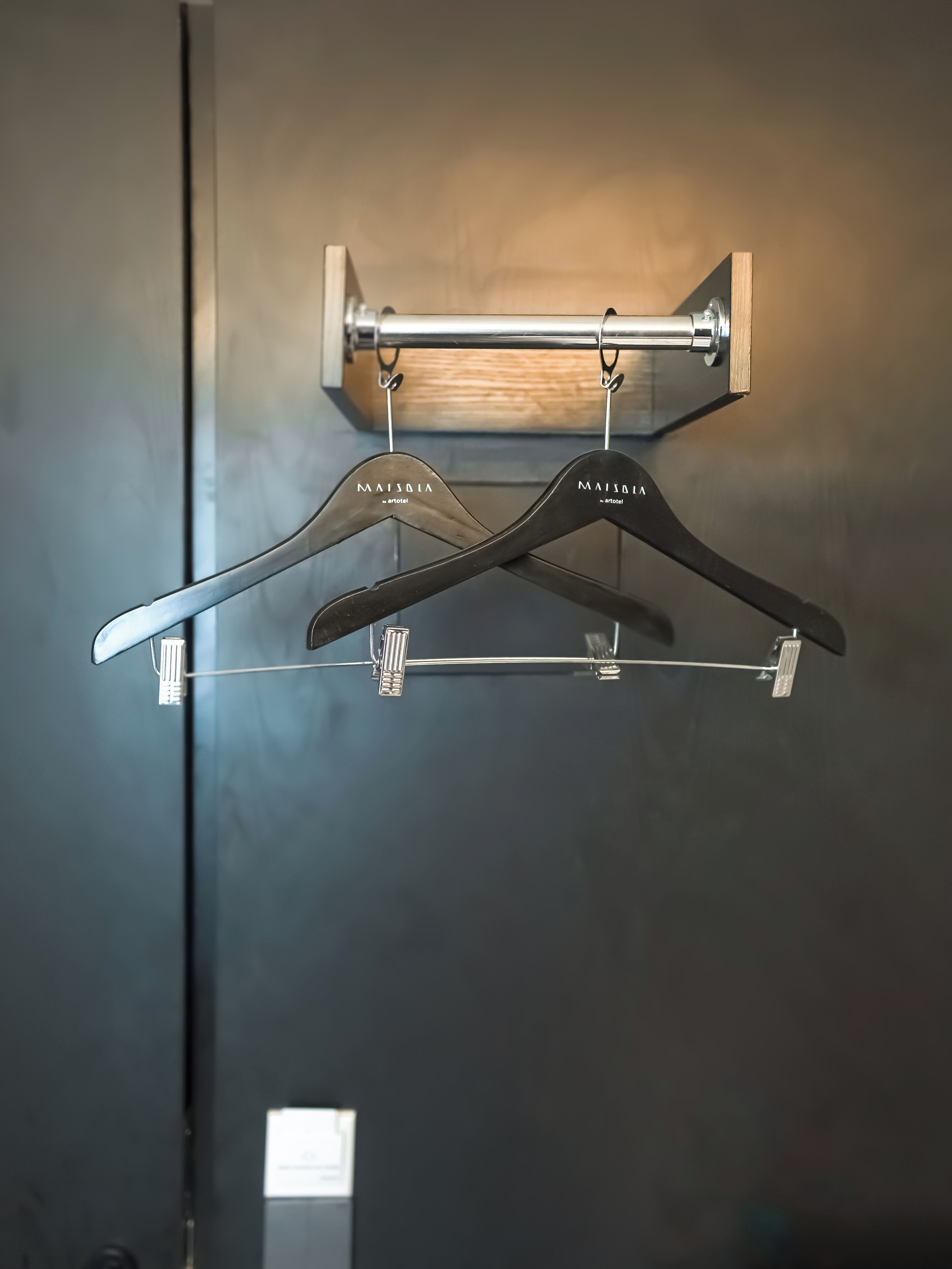 A wall-mounted metal rack with two black hangers labeled "MAISBIA" and "artotel" against a dark wall.