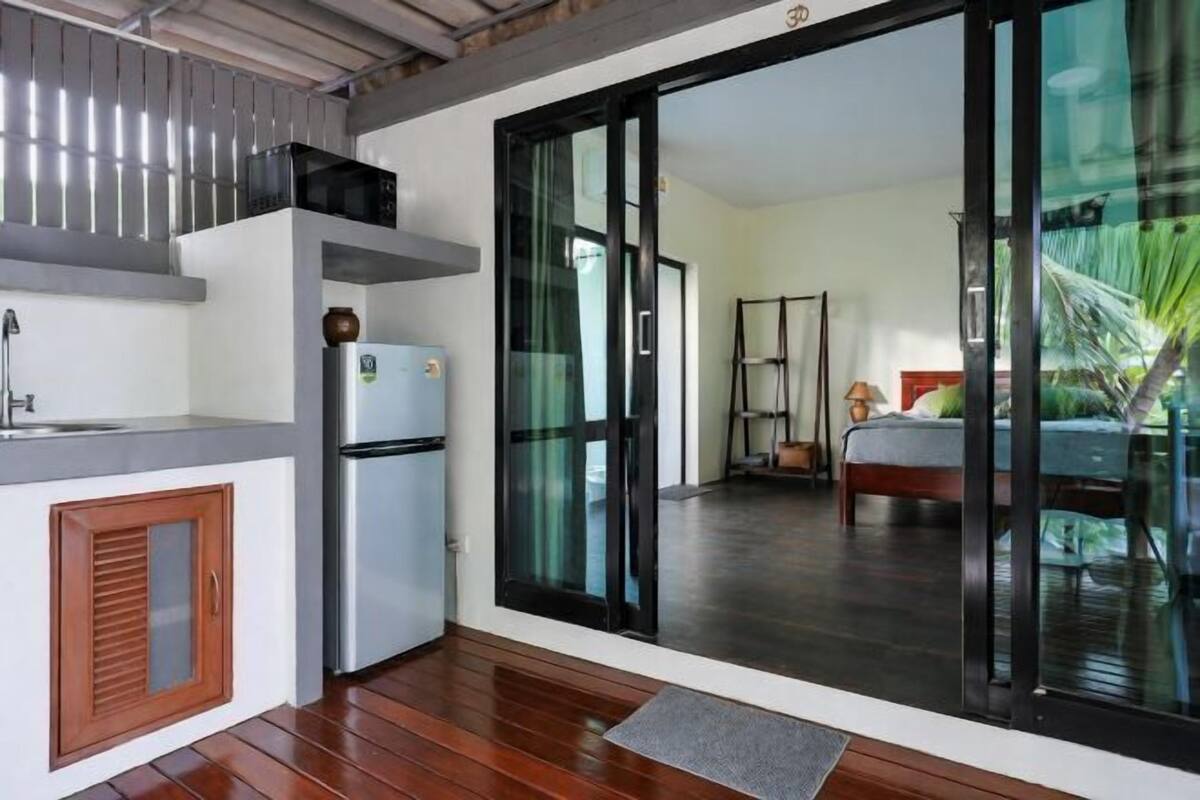 A compact kitchen with a refrigerator, a sink, and a window overlooking a bedroom with a bed and a nightstand.