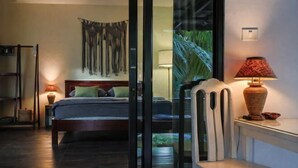 A hotel room with a bed, a chair, a lamp, a wall-mounted shelf, and a view through a glass door. - Island’s Gift – Private Design House for 2 Near Sunset Beach (Koh Phangan)
