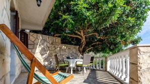 A patio with a wooden chair, a table, and chairs, surrounded by a stone wall and a large tree. - Apartments Katica's Place - One Bedroom Apartment With Terrace and City View (Blato)