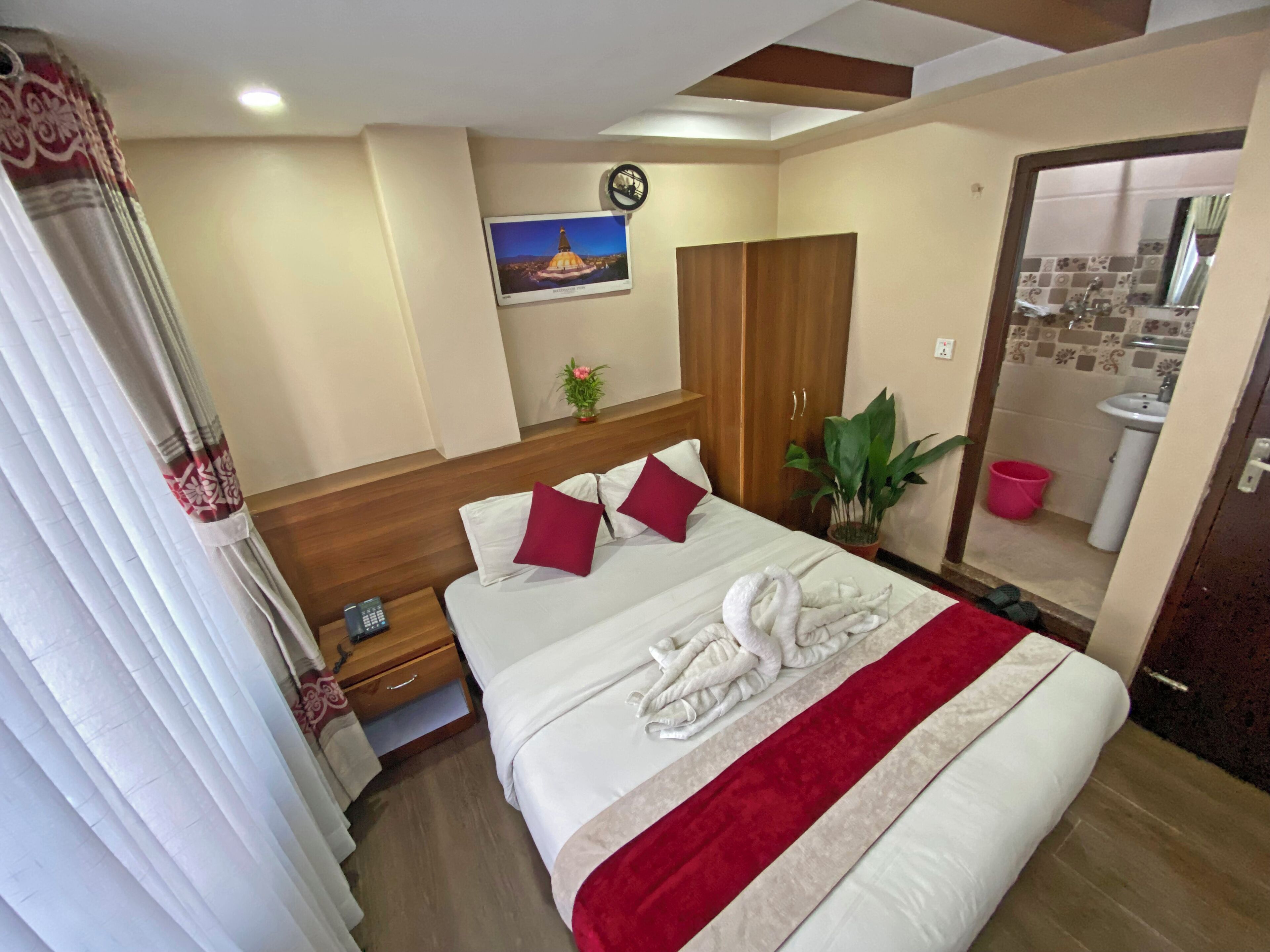 A hotel room with a neatly made bed, a wooden headboard, a clock, a potted plant, and a bathroom visible through an open door.