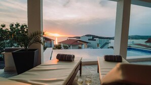 Una terraza iluminada por el sol con vistas a la puesta de sol sobre un pueblo costero, con dos sillones y una mesa con vasos.