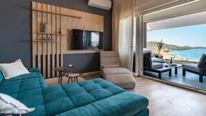 Una habitación de hotel moderna con una cama grande, un televisor montado en la pared, una mesita pequeña y un sofá. La habitación tiene un gran ventanal corredero que da a un balcón con vistas al mar.
