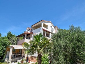 Un edificio a più piani con balconi, un portico coperto e vegetazione circostante.