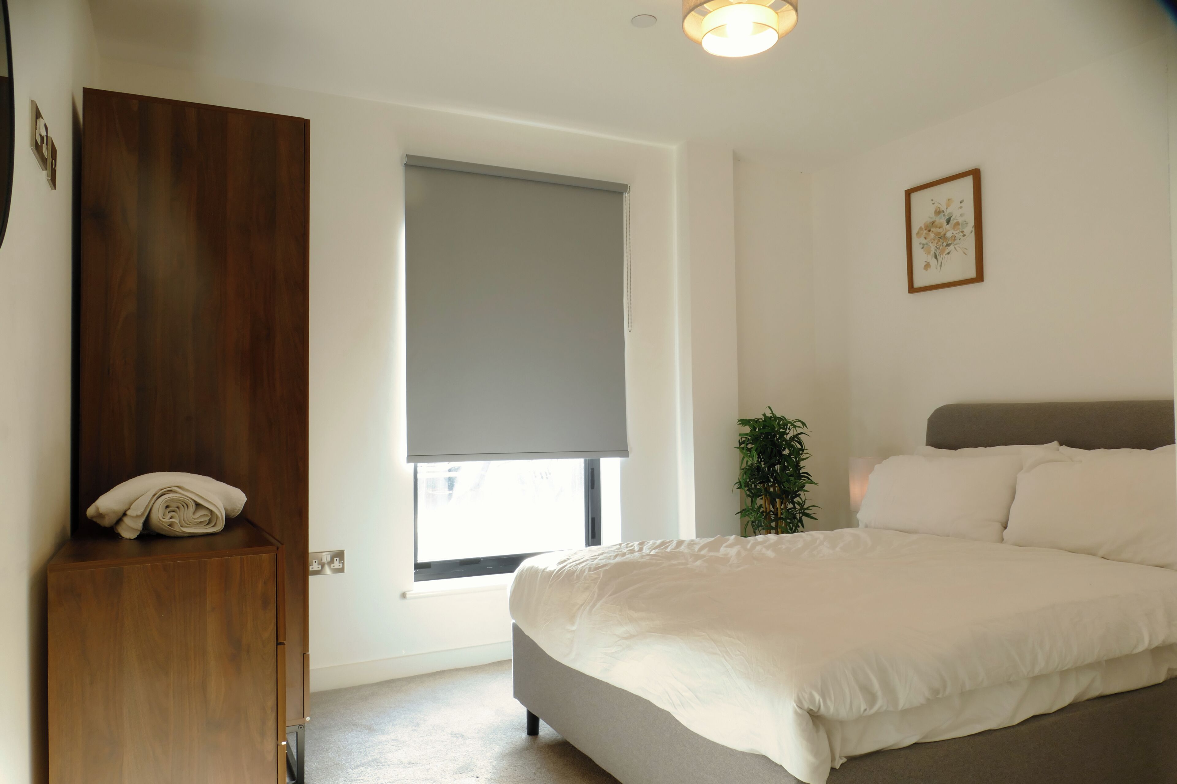 A bedroom with a bed, a wooden wardrobe, a window with a blind, and a small plant.
