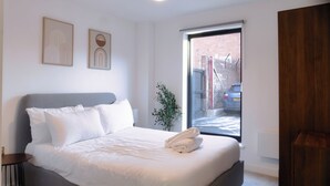 Ein modernes Schlafzimmer mit einem großen Bett, einem Nachttisch, einer Topfpflanze und Blick auf die Straße durch ein Fenster.